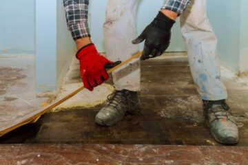 A construction worker is removing flooring from the floors of a home while wearing appropriate safety gear.
