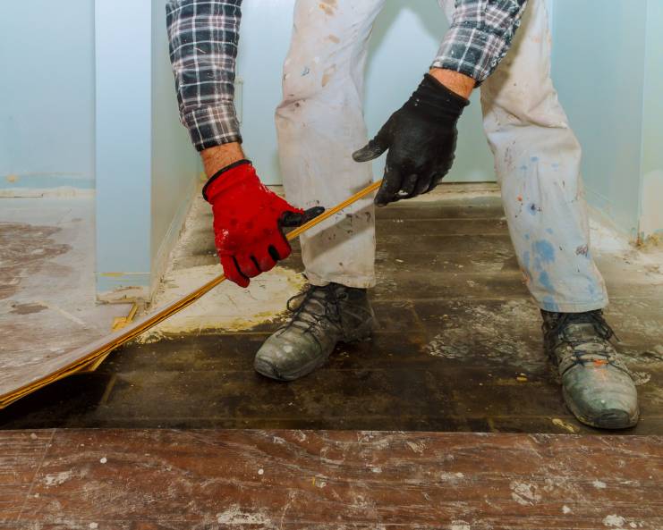 A construction worker is removing flooring from the floors of a home while wearing appropriate safety gear.