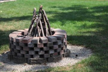 A round fire pit made out of bricks standing in a lush green backyard. A pyramid of sticks is coming up from the center.