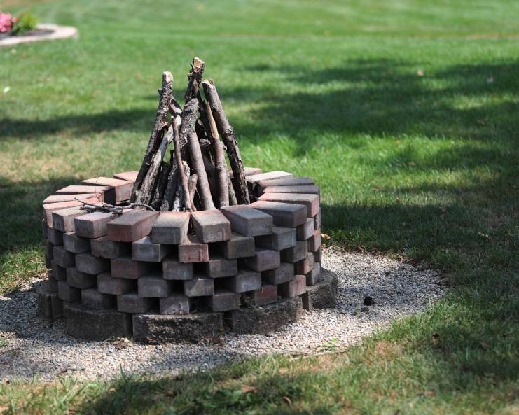 A round fire pit made out of bricks standing in a lush green backyard. A pyramid of sticks is coming up from the center.