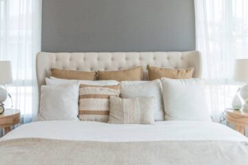 A cream headboard sits between two windows. The bed has several tan and cream-colored throw pillows, along with a blanket.