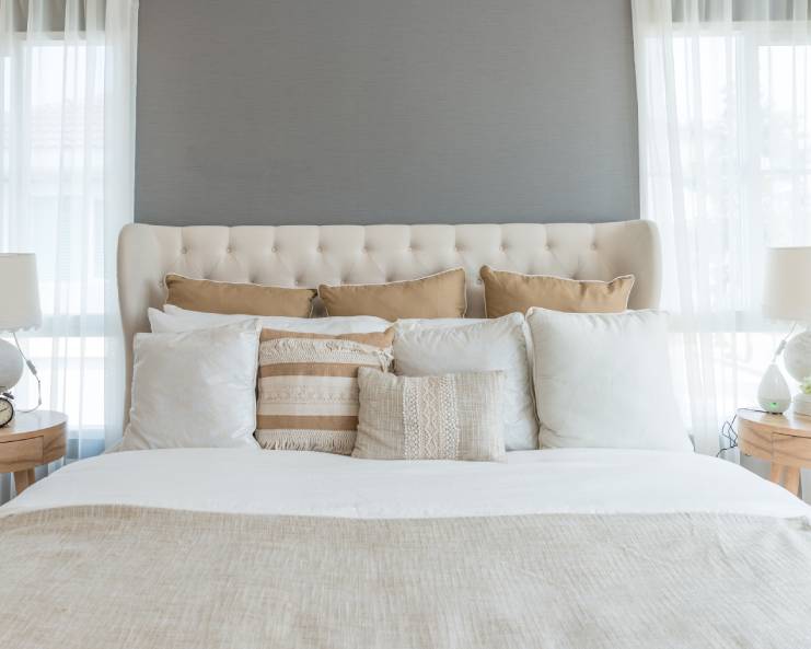 A cream headboard sits between two windows. The bed has several tan and cream-colored throw pillows, along with a blanket.