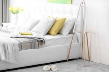 Cozy slippers sit on the gray carpeted floor of a soothing white bedroom. A vase of yellow tulips sits on the side table.