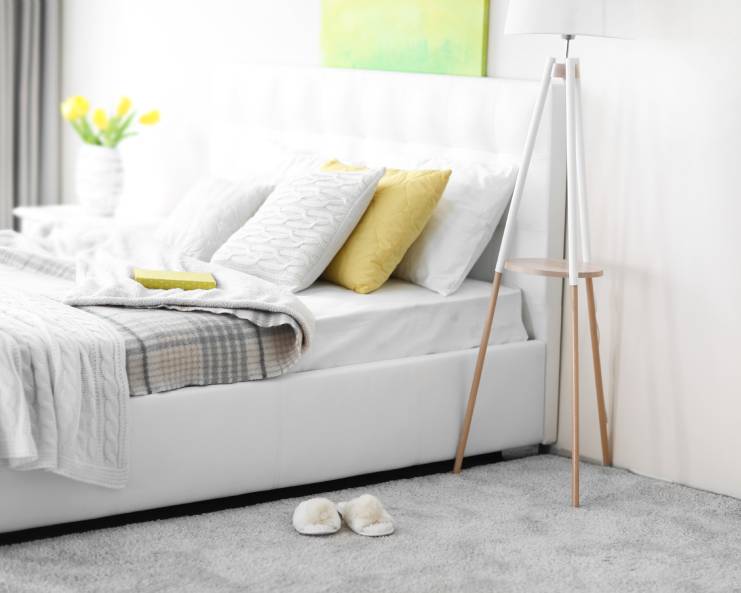 Cozy slippers sit on the gray carpeted floor of a soothing white bedroom. A vase of yellow tulips sits on the side table.