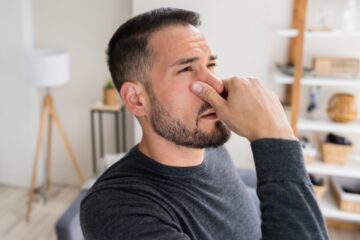 A man holding his nose tightly and looking frustrated because of a bad odor that he smells inside his house.