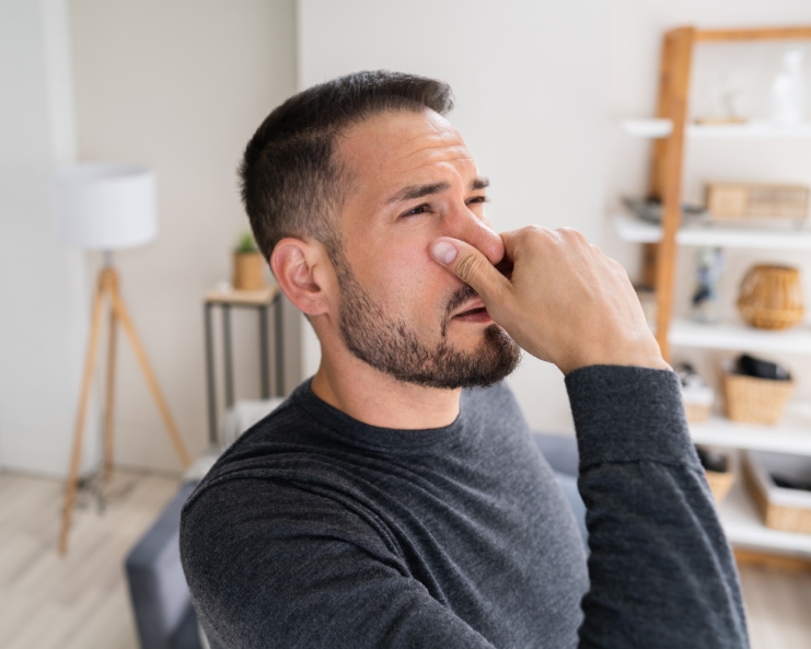 A man holding his nose tightly and looking frustrated because of a bad odor that he smells inside his house.