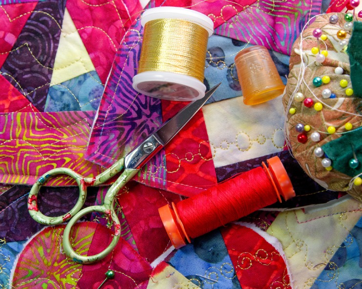 A red and gold thread spool sits next to fabric scissors and pins on top of brightly colored fabric.