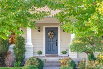 Large, mature trees surround the front porch and front door of a home. The front door is a rich blue color.