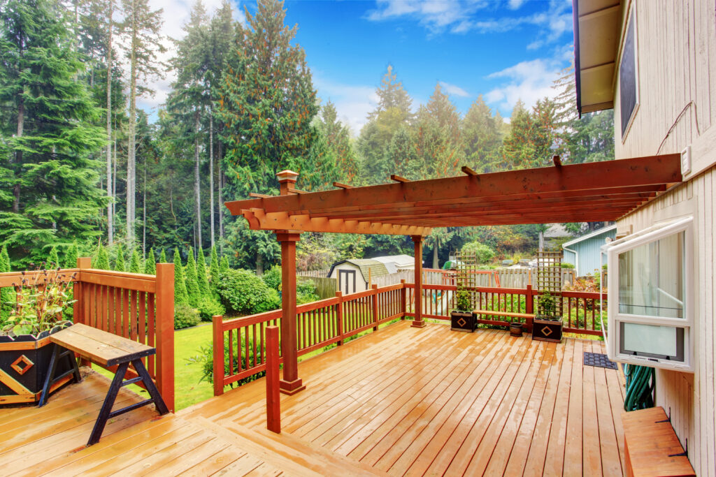 An outdoor wooden walkout deck with an attached pergola after a rainy afternoon. The deck has potted plants and a bench.