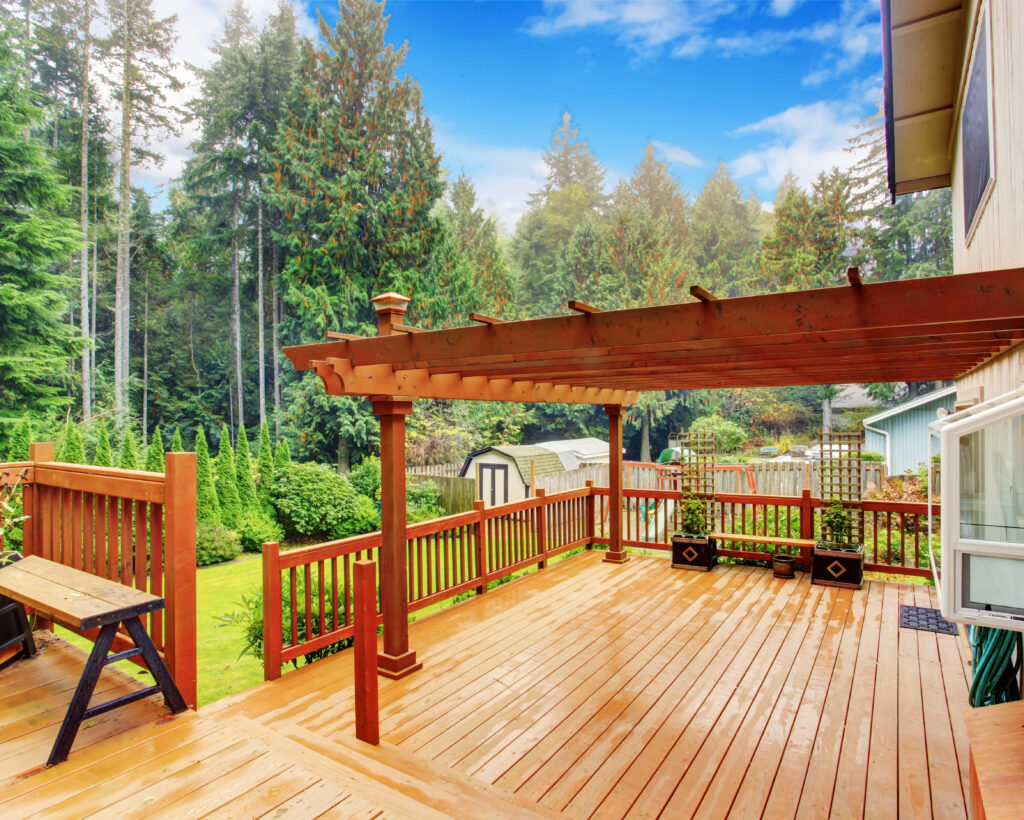 An outdoor wooden walkout deck with an attached pergola after a rainy afternoon. The deck has potted plants and a bench.