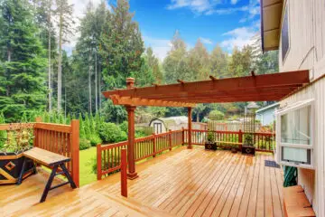 An outdoor wooden walkout deck with an attached pergola after a rainy afternoon. The deck has potted plants and a bench.