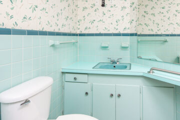 An outdated bathroom features blue tiles and a wallpaper with blue flowers. The bathroom vanity matches the tile.