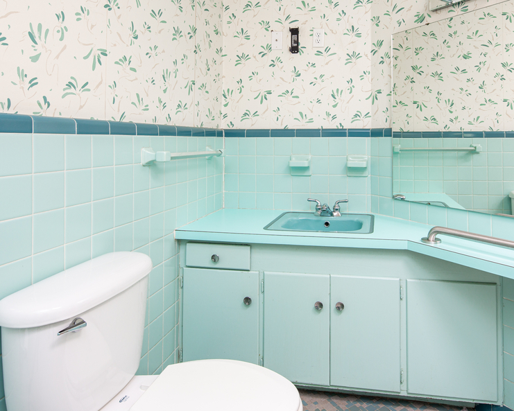 An outdated bathroom features blue tiles and a wallpaper with blue flowers. The bathroom vanity matches the tile.