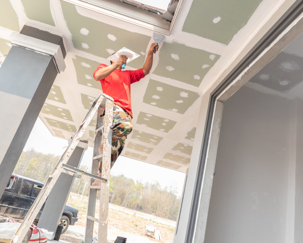 A construction worker with an orange shirt standing on a ladder, carefully applying plaster to a ceiling with a trowel.