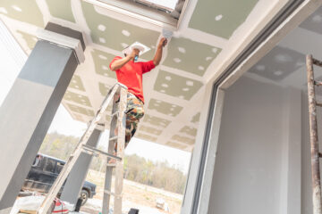 A construction worker with an orange shirt standing on a ladder, carefully applying plaster to a ceiling with a trowel.