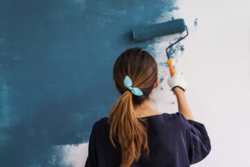 A woman in a blue shirt uses a roller to paint the walls a deep teal color. Her hair is in a ponytail with a bow.