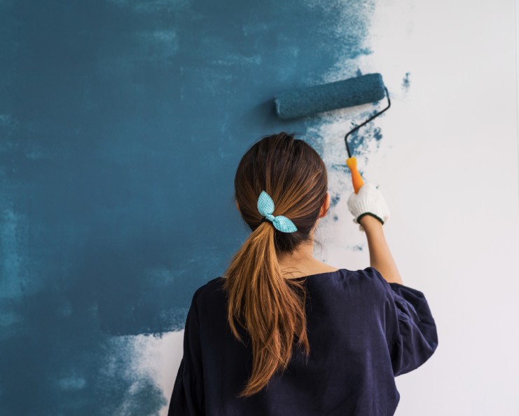 A woman in a blue shirt uses a roller to paint the walls a deep teal color. Her hair is in a ponytail with a bow.