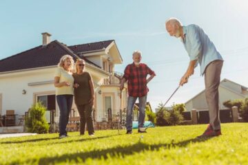 Four older adults gather in a backyard on a sunny afternoon as one man prepares to swing his club and putt a golf ball.