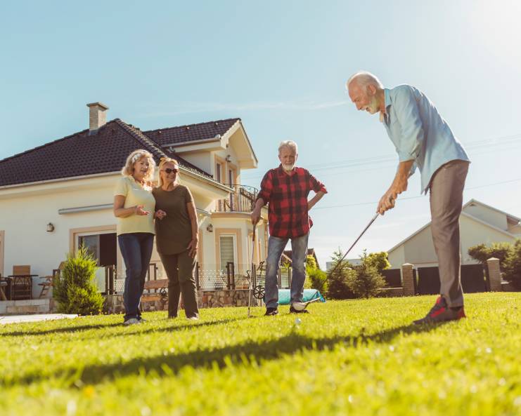 Four older adults gather in a backyard on a sunny afternoon as one man prepares to swing his club and putt a golf ball.