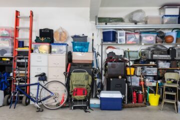 A garage with a concrete floor and white walls features household items such as filing cabinets, a ladder, and a bicycle.