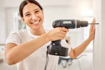 A woman in a striped shirt smiling as she drills a hole with a black power drill into a cream-colored wall.