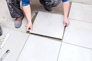 A person wearing a blue shirt and dirty, black pants is installing white tiles on the bathroom floor.