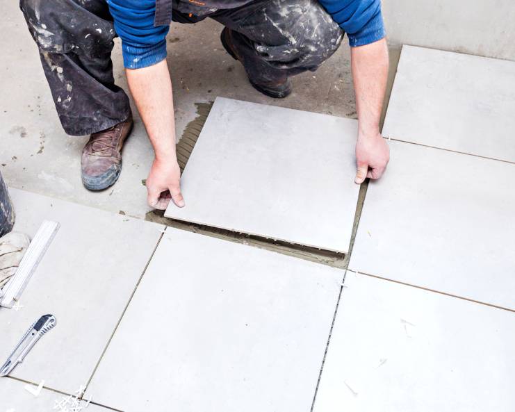 A person wearing a blue shirt and dirty, black pants is installing white tiles on the bathroom floor.
