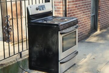 An old household stove sitting outside on the sidewalk. It has a stainless steel door and drawer with black accents.