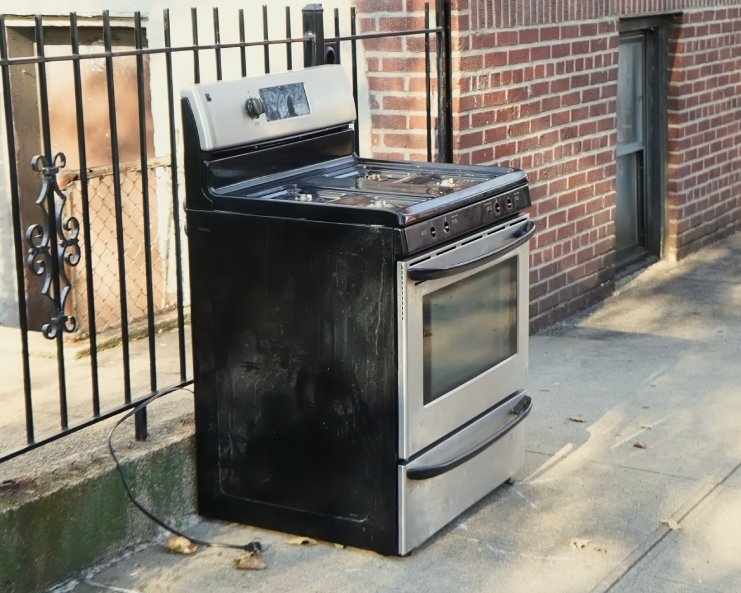 An old household stove sitting outside on the sidewalk. It has a stainless steel door and drawer with black accents.