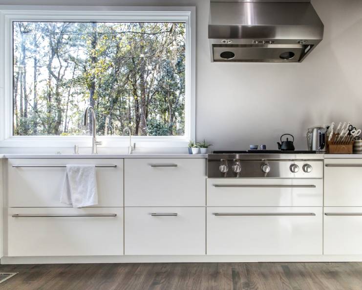 A modernly designed kitchen interior with white cabinets and a large picture window above the sink.