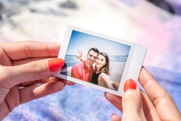 Two hands holding a polaroid photo of a couple at the beach. The couple is standing close to the shoreline and smiling.