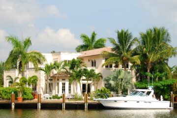 A large home sits at the edge near a body of water in between several palm trees. There is a white yacht docked in front.