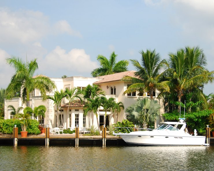 A large home sits at the edge near a body of water in between several palm trees. There is a white yacht docked in front.