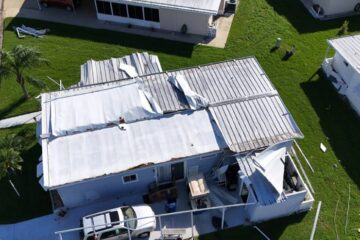 An aerial view of a damaged mobile home roof. The green grass yard surrounding the home is scattered with debris.