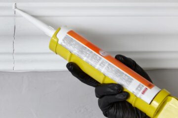 A person wearing black gloves uses a caulk gun to seal a crack in a piece of wood trim inside of a home.