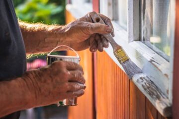 A man using a paintbrush to apply white paint to the trim of a window on the side of a wooden building.