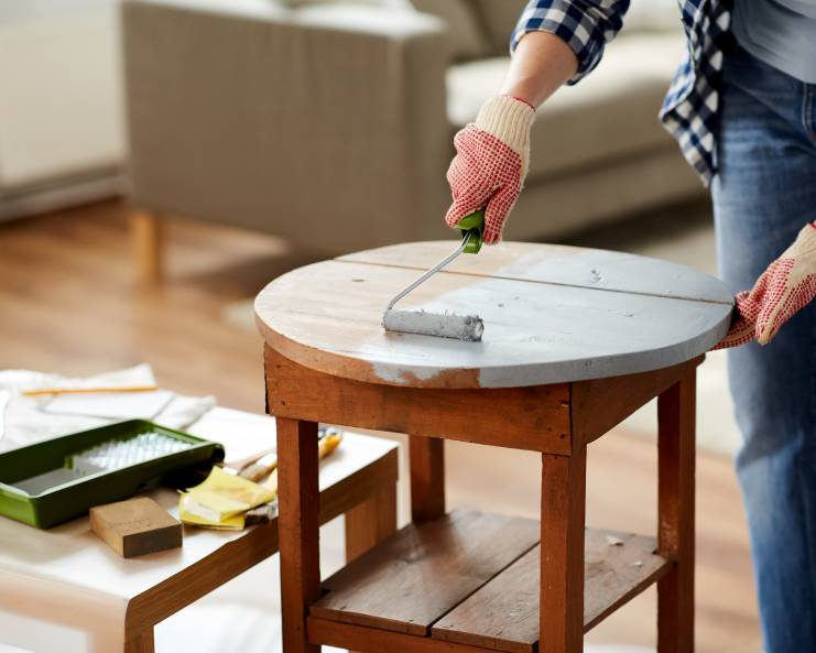 A person wearing gloves uses a small paint roller to paint the round top of a wooden table in a home setting.