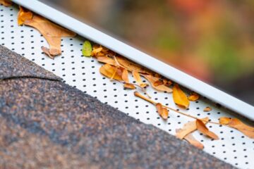 A gutter with a mesh gutter guard extending from the edge of an asphalt-shingled roof. There are leaves in the gutter.