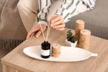 A person with a green and white striped shirt reaches for a reed in a jar from a reed diffuser set on a wooden table.