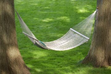 A white hammock made of thick strings hanging between the trunks of two trees with a blue pillow on one side.