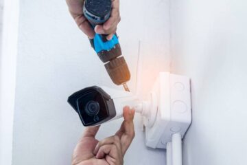 A person installing a home camera in the corner of a room. They are holding a drill and positioning the camera in place.