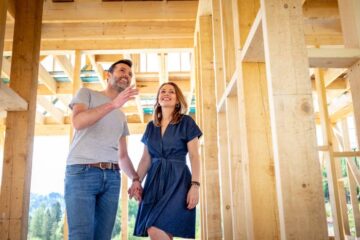 A man and women holding hands and smiling as they stand inside of a wood building frame; the sun shines in the background.