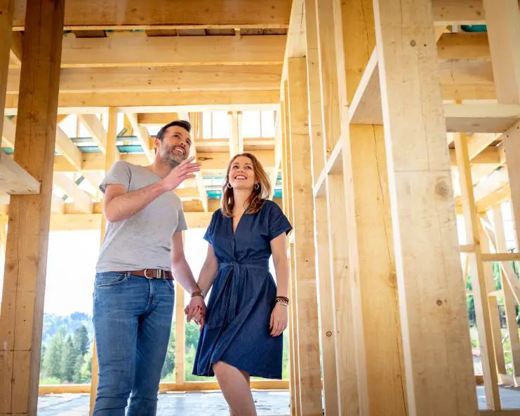 A man and women holding hands and smiling as they stand inside of a wood building frame; the sun shines in the background.