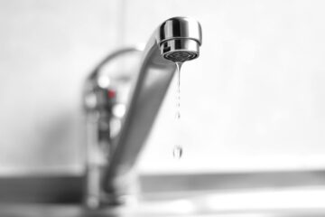 A stainless steel faucet leaking water from its tap. The background is blurry, and the base of the faucet is blurred.