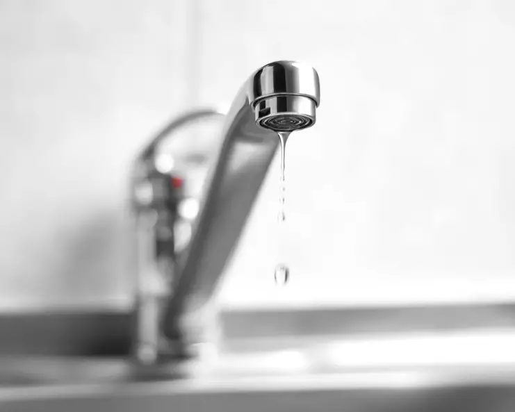 A stainless steel faucet leaking water from its tap. The background is blurry, and the base of the faucet is blurred.