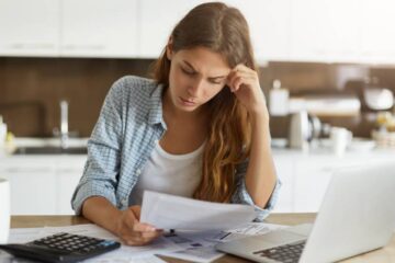 A woman sitting at a table with a laptop, calculator and paperwork. She rests her hand on her head as she looks at a paper.