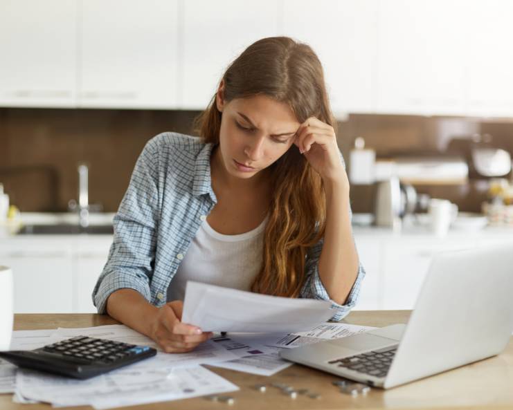 A woman sitting at a table with a laptop, calculator and paperwork. She rests her hand on her head as she looks at a paper.