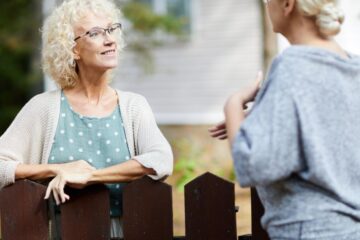 Two mature women who are neighbors speaking to each other are separated by a dark brown wooden picket fence.