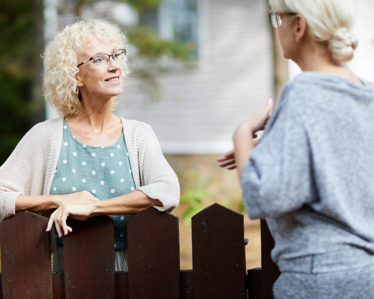 Two mature women who are neighbors speaking to each other are separated by a dark brown wooden picket fence.