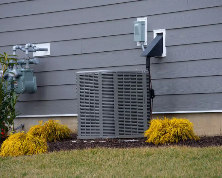 An air conditioner installed on a patch of dirt and concrete outside a home with gray siding and yellow bushes.